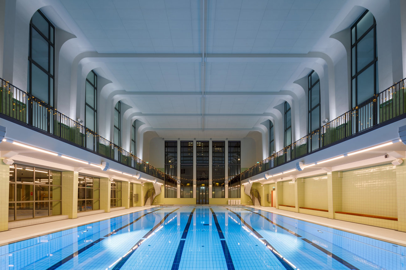 Inomhussimhall med simbassäng, banindelare, höga fönster och galleri.