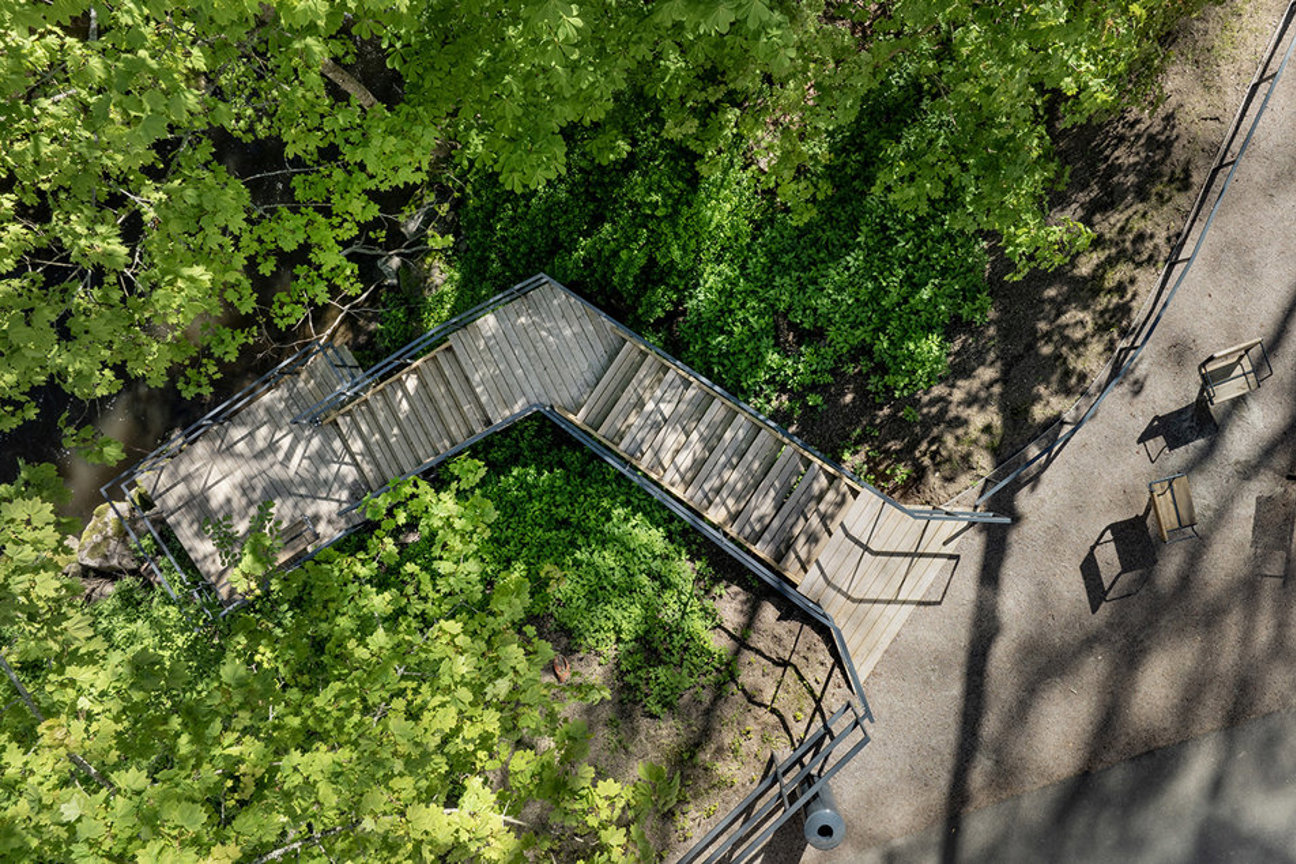 Luftbild av träbro med räcken omgiven av gröna träd och buskar, med stolar på en grusväg.