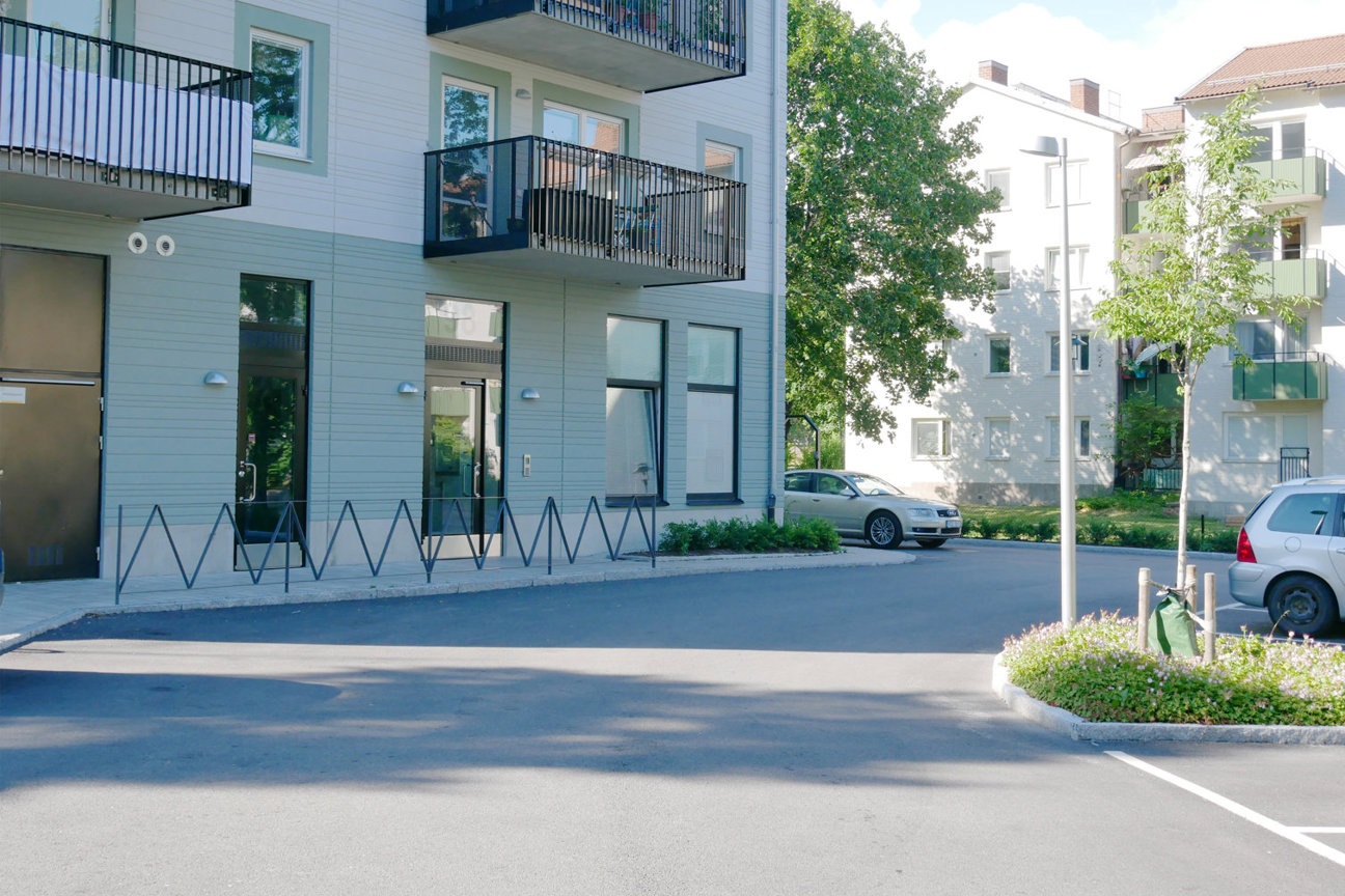 Ljusblå bostadshus med balkonger, parkerade bilar och träd vid en gata.