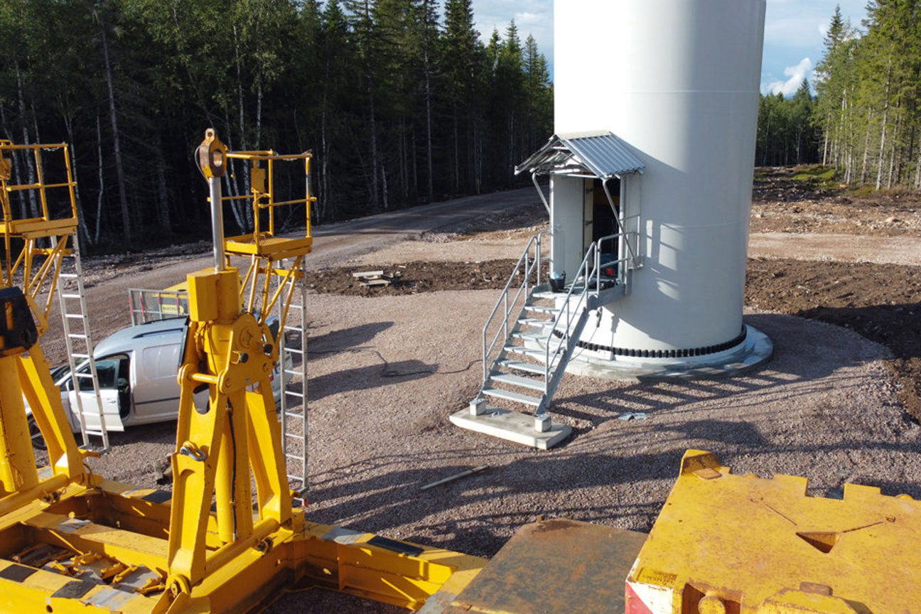 Vindkraftverk med gula byggmaskiner, vit bil och trappa på grusbeläggning omgiven av skog.