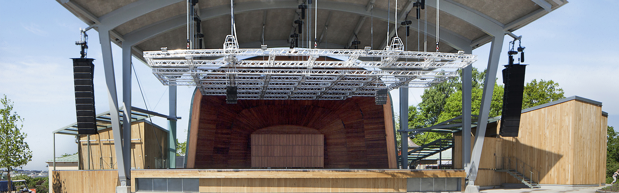 Utomhus-scen med träfasad, metallbalkar och högtalare under en takkonstruktion. Träd och byggnader i bakgrunden.