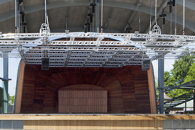 Utomhus-scen med träfasad, metallbalkar och högtalare under en takkonstruktion. Träd och byggnader i bakgrunden.