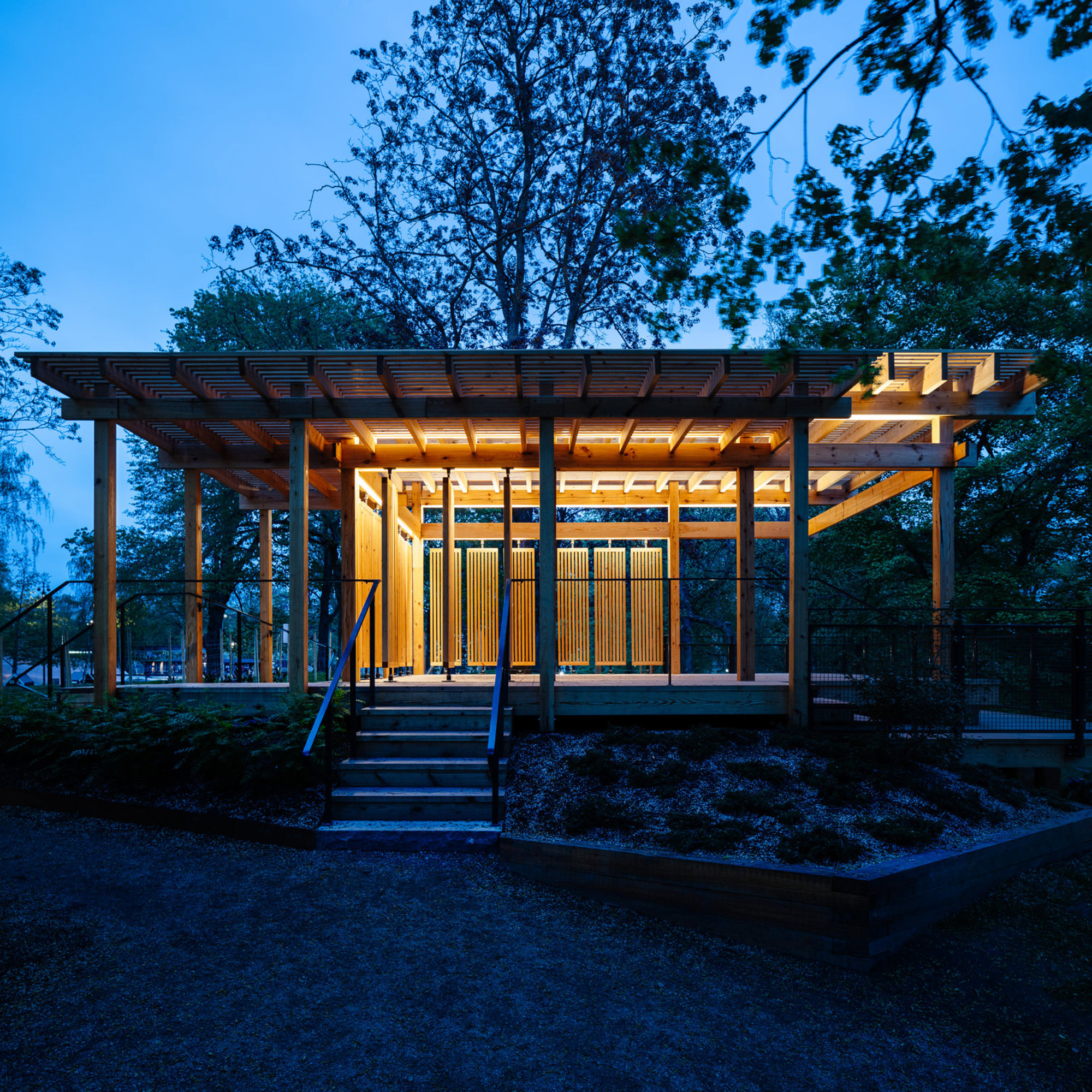 En träpaviljong med trappa och belysning omgiven av träd vid skymning.