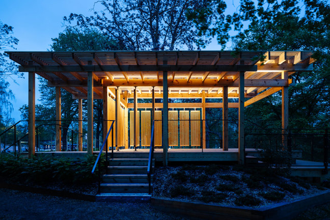 En träpaviljong med trappa och belysning omgiven av träd vid skymning.