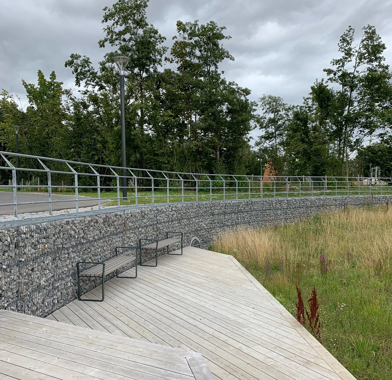 Trädgårdsplats med träbänkar, stenvägg och metallräcke under molnigt väder.