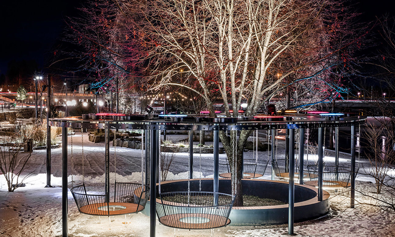 Nattbild av en trädgård med cirkulära svängbänkar, snö och belysning runt ett träd.