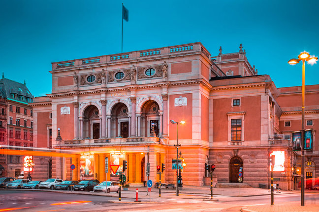 Stor historisk byggnad med arkader och gatubelysning vid kvällstid.	
