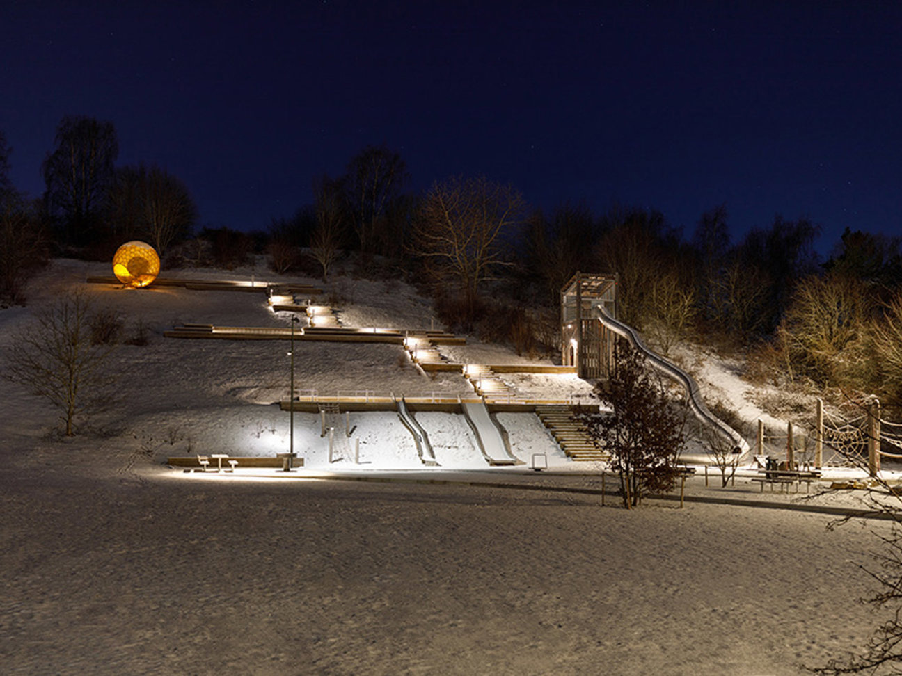 En snöig lekplats på natten med belysta rutschbanor och en stor sfärisk struktur.