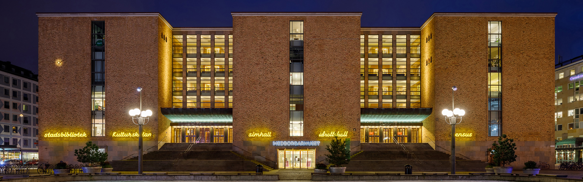 Nattbild av en tegelbyggnad med belysta fönster och neonreklamer för stadsbibliotek och Kulturskolan