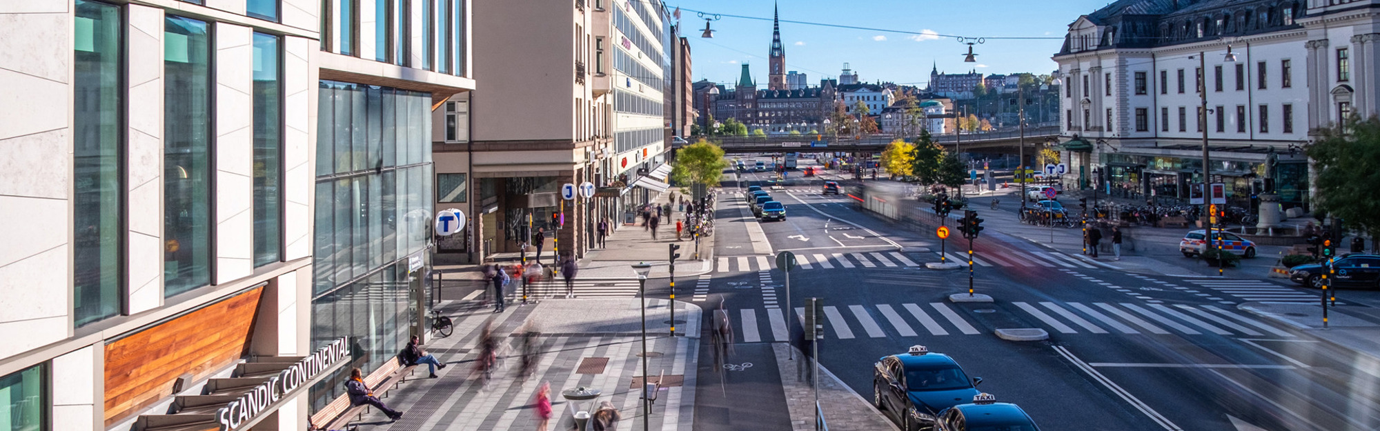 Stadsgata med byggnader, bilar och gående.