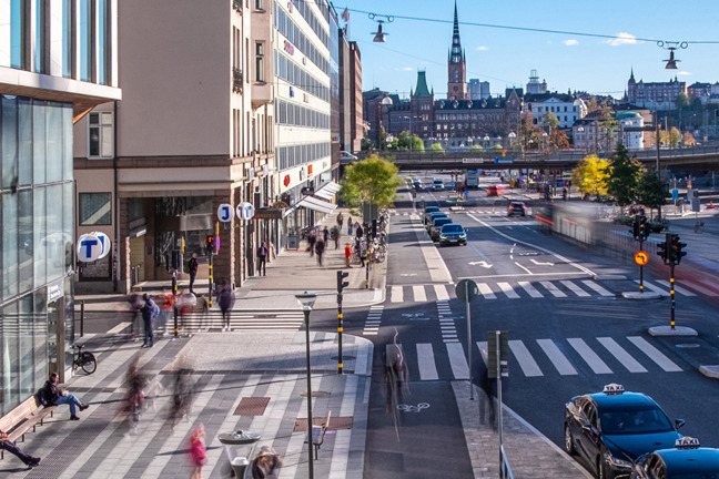 Stadsgata med byggnader, bilar och gående.