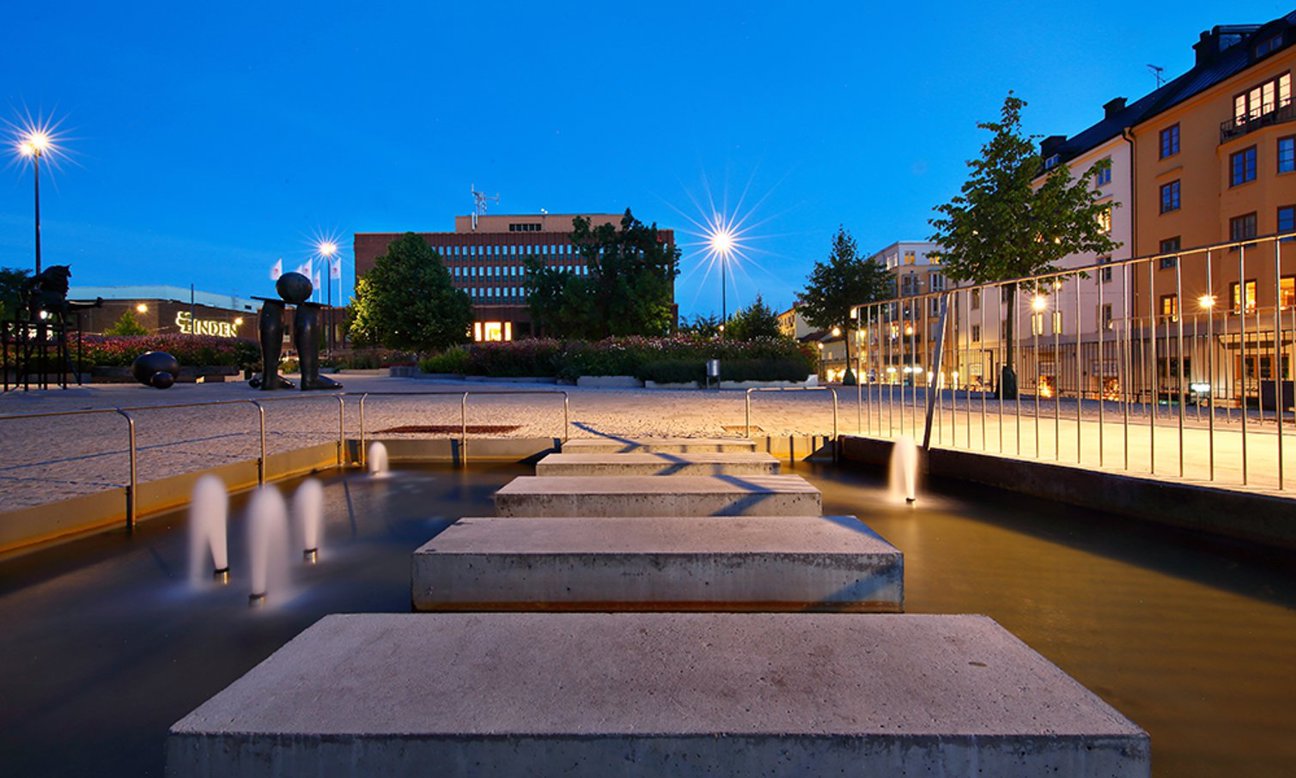 En kvällsutsikt över en stadsplats med betongplattformar, springbrunnar, gatubelysning, abstrakta skulpturer och byggnader mot en blå himmel.