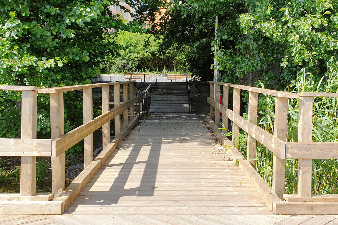 En träbro med räcken omgiven av gröna träd och buskar, med trappor i bakgrunden.