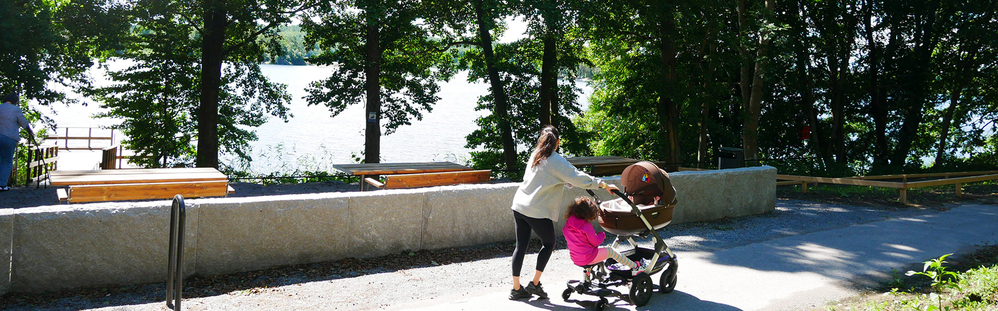 En kvinna och ett barn vid en barnvagn på en promenadväg vid en sjö med bänkar och träd.