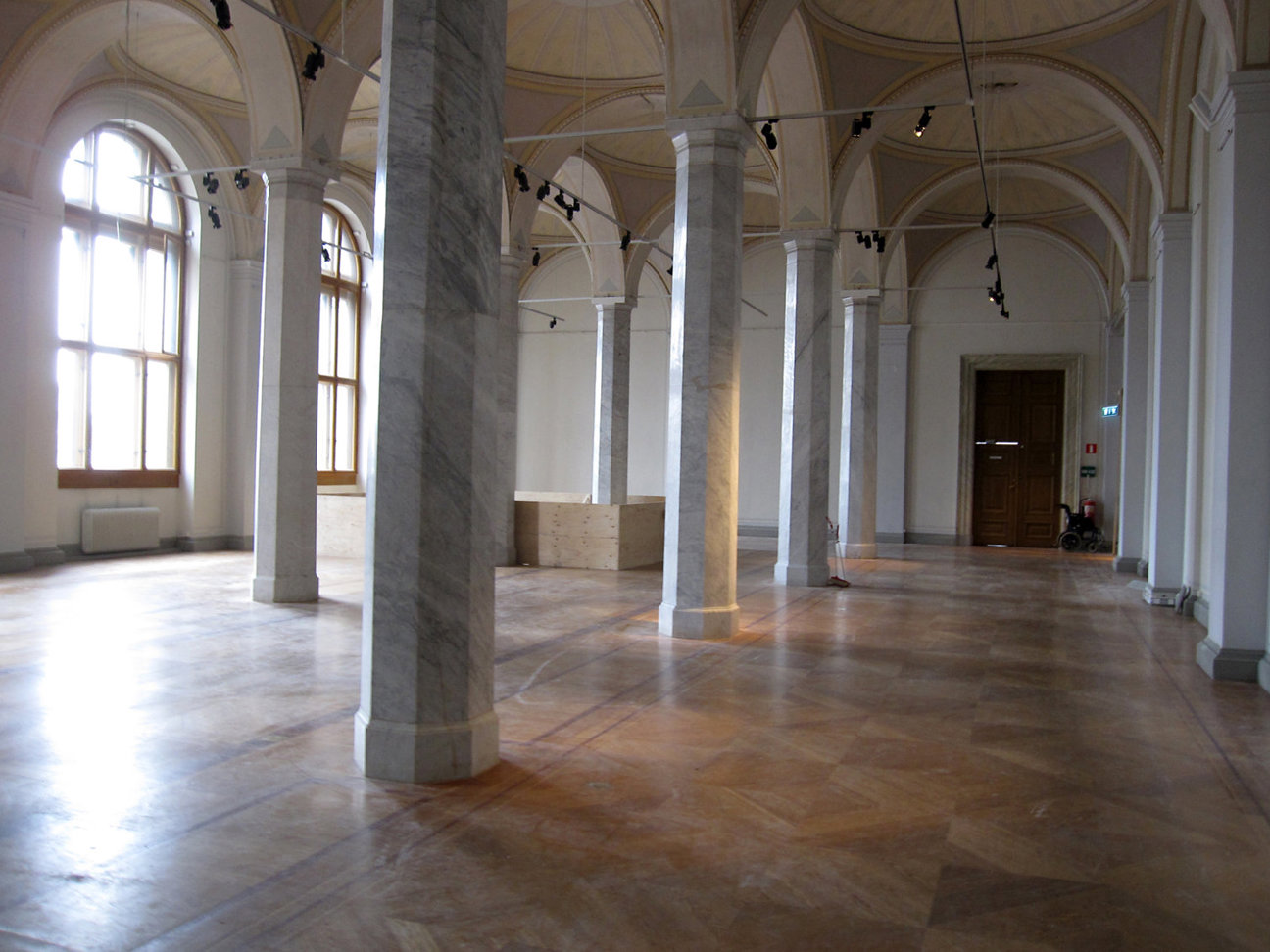 Stort rum med höga valv, pelare, stora fönster och trägolv. Rullstol vid dörr.