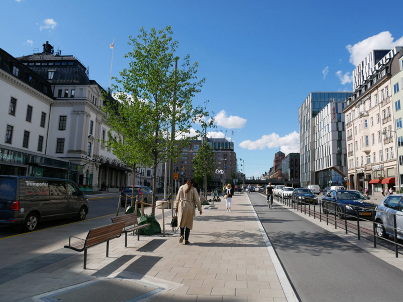 Gata med byggnader, gångbana, träd, parkerade bilar och gående människor under klar himmel.