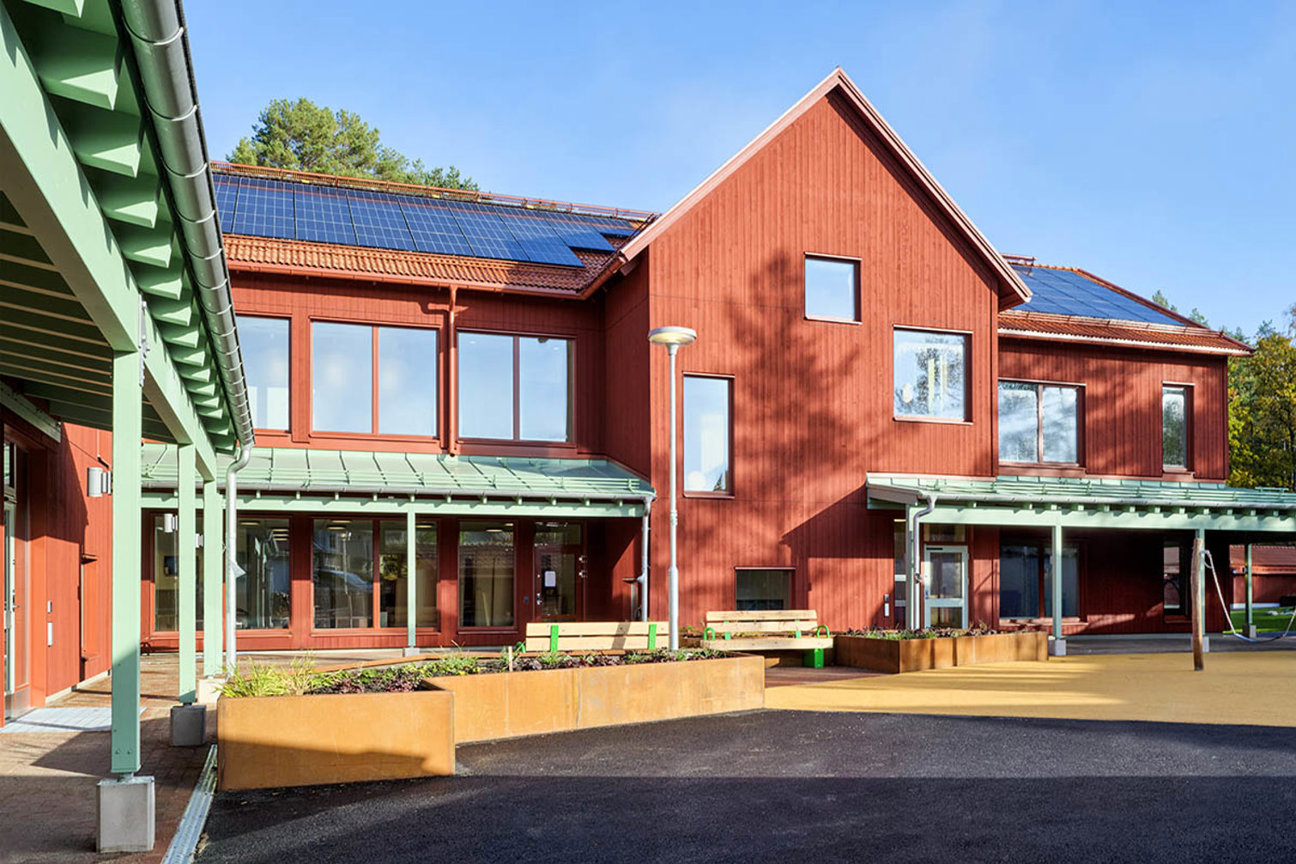 Rött trähus med solpaneler och gröna taklängor, planteringskasser och bänkar framför byggnaden.