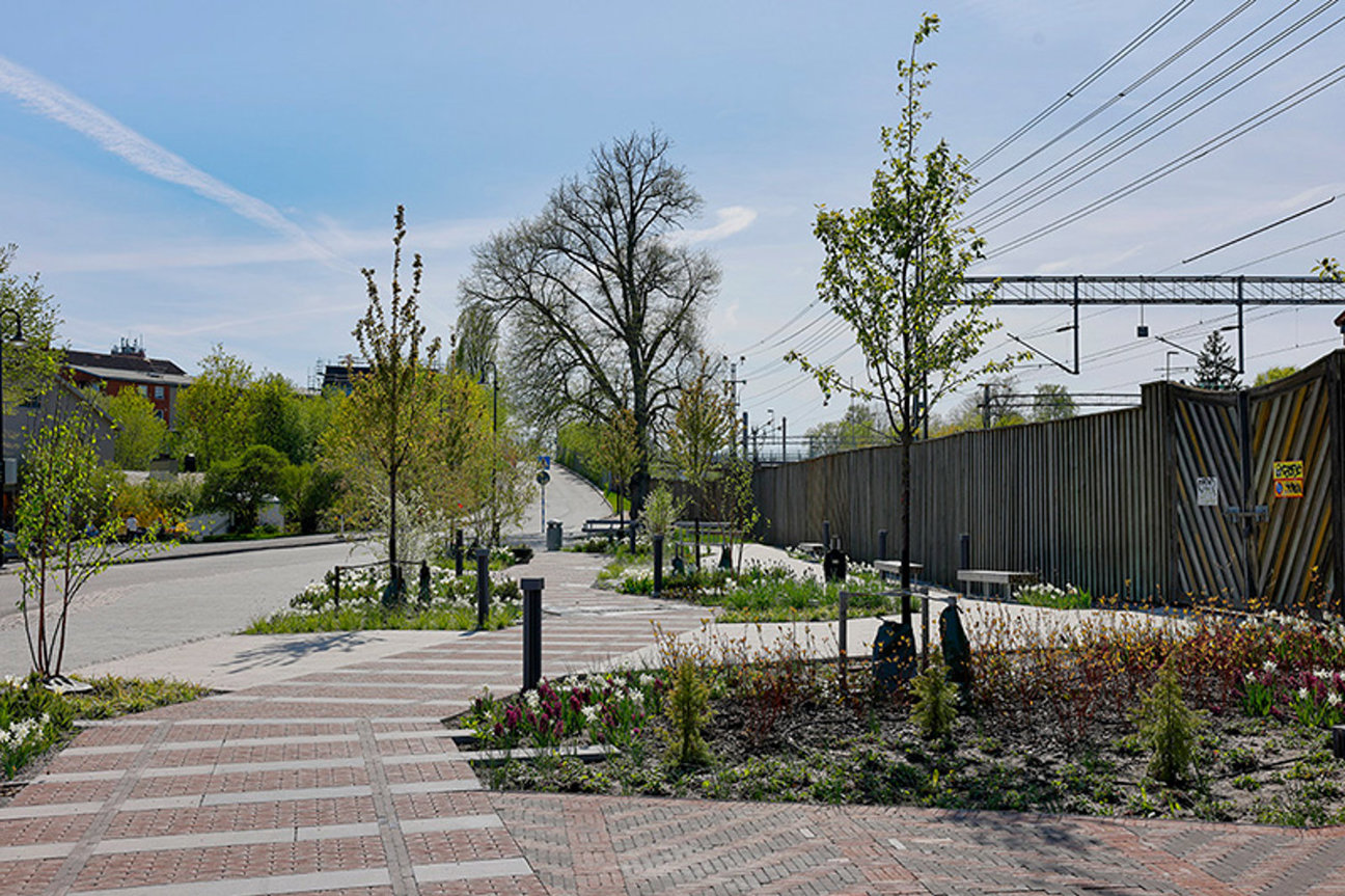 Stenlagd gångväg med blommor, träd och staket vid järnvägsspår under blå himmel.