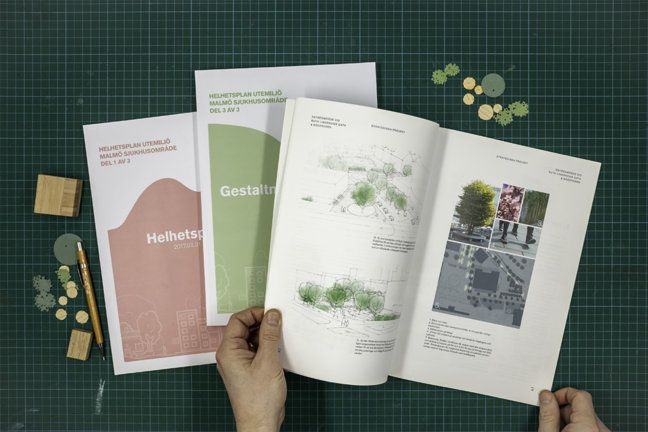Dokument för helhetsplan Malmö sjukhusområde med skisser och bilder. Två händer håller öppen dokument på grön rutig underlägg.