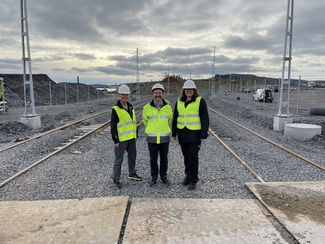 Tre personer i gula reflexvästar och vita hälmar står på en grusad yta med järnvägsspår.