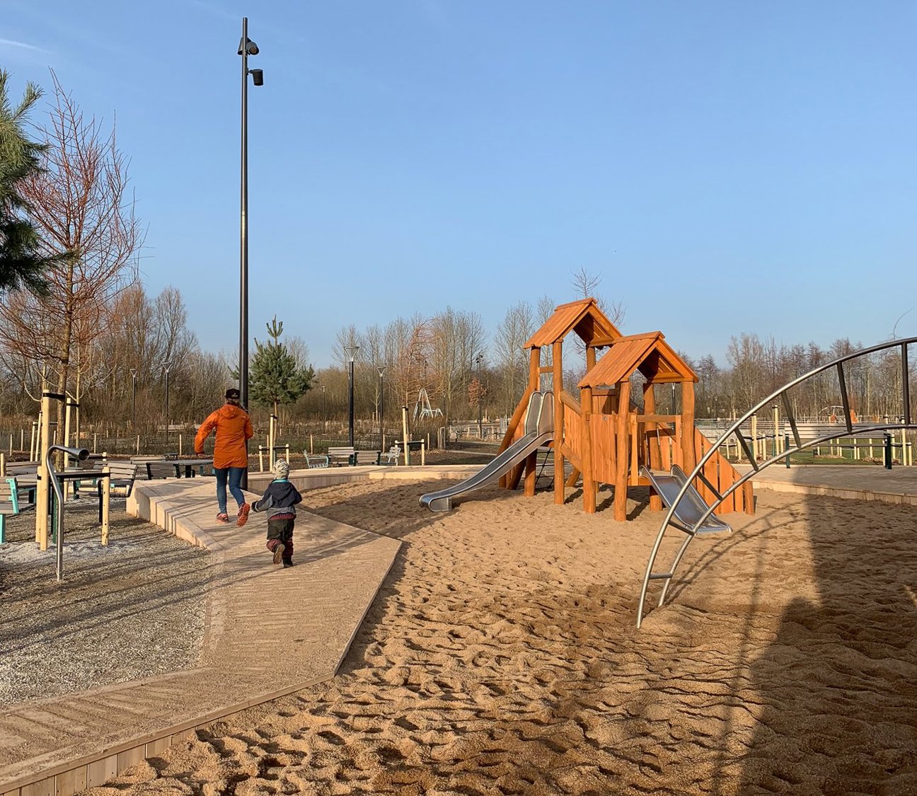 Lekplats med trästruktur, rutschbanor och sand. Två personer går på gångväg.