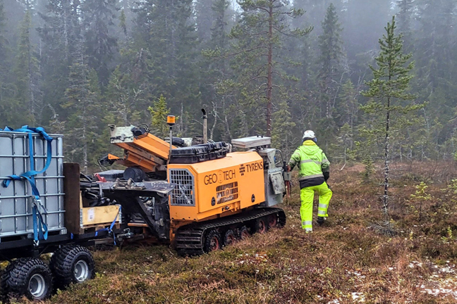Person i lysande grön arbetskläder bredvid maskin och behållare i skog.