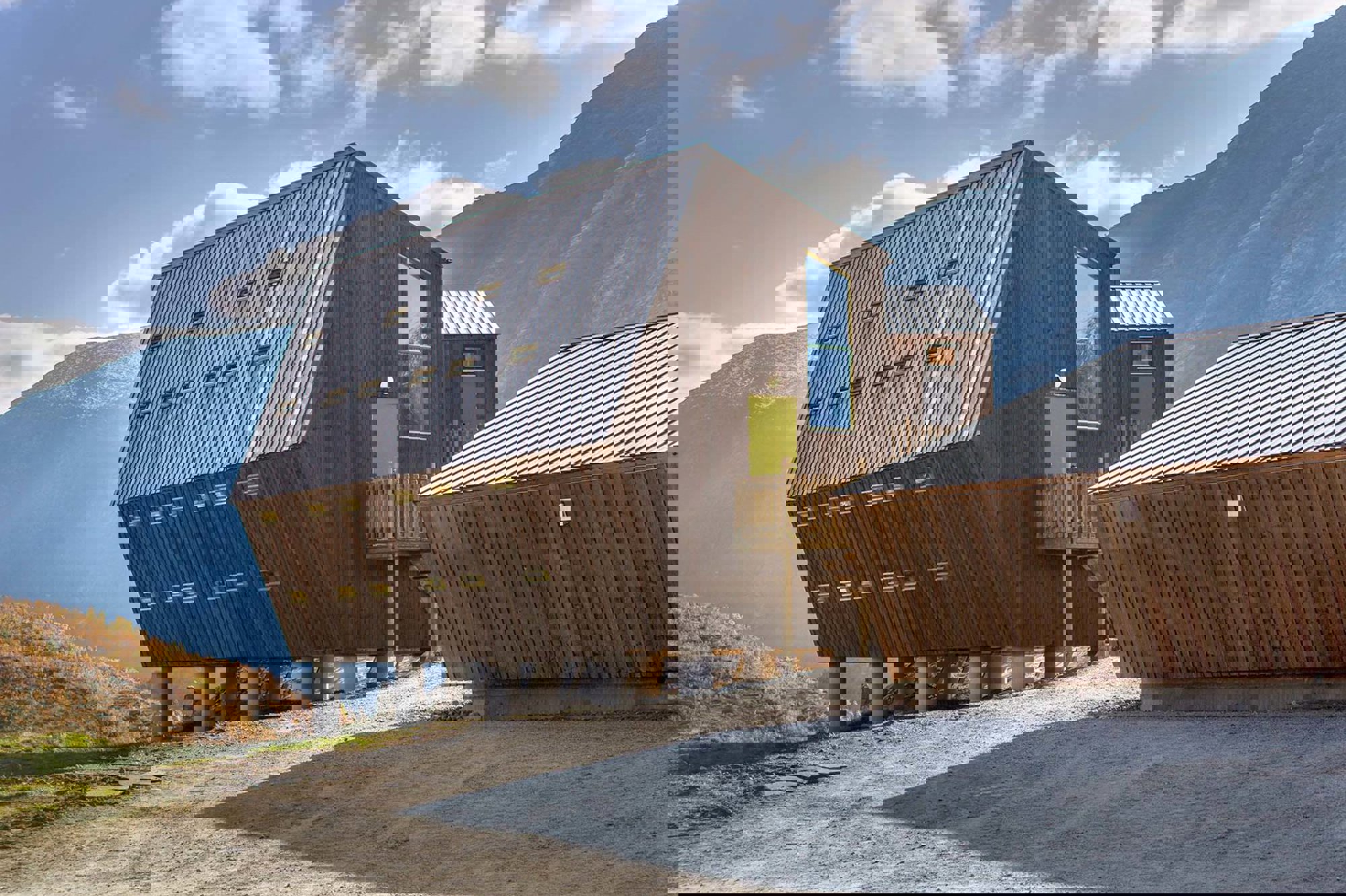 Modernt trähus med lutande metalltak och flera små fönster, beläget i bergigt landskap under blå himmel.