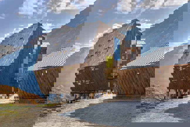 Modernt trähus med lutande metalltak och flera små fönster, beläget i bergigt landskap under blå himmel.