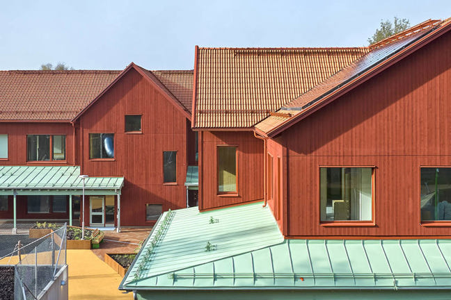 Rött trähus med gröna tak omgivet av träd och lekplats.