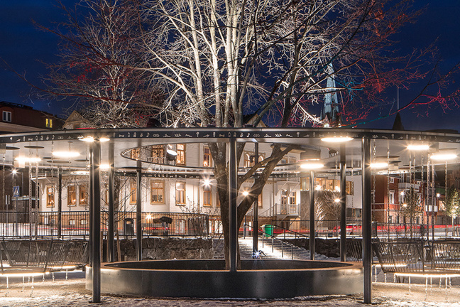 Nattlig torgplats med snöig mark, belysta byggnader, ett blottat träd och en cirkulär ljusstruktur.