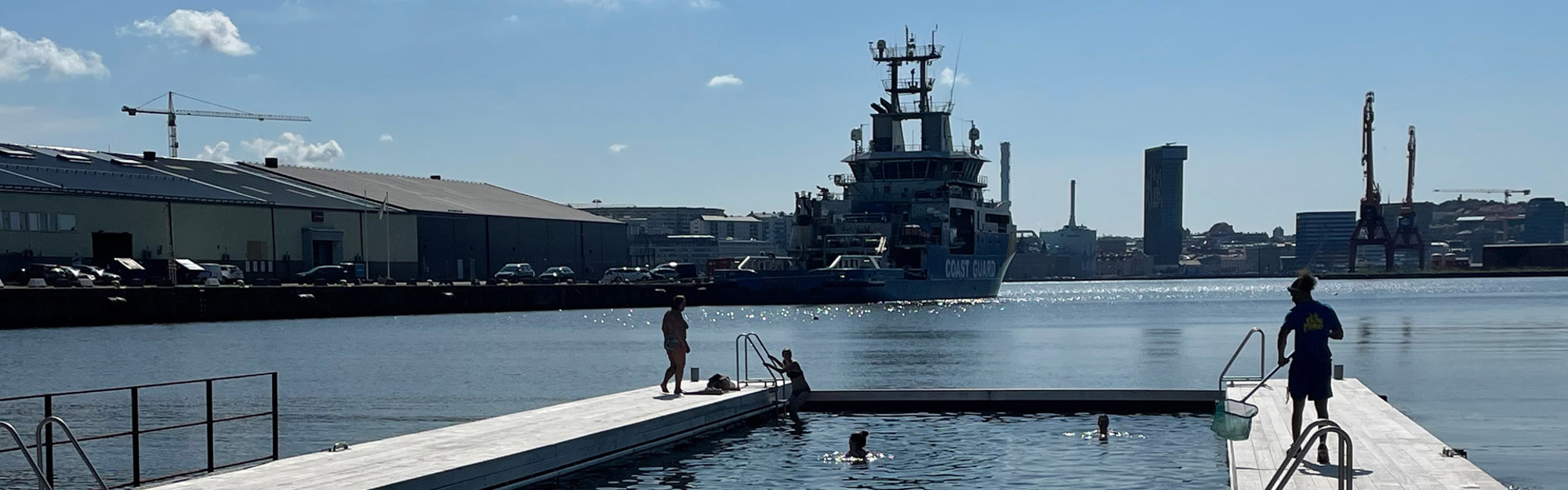 Personer på brygga och i vatten, kustbevakningsfartyg och kranar i hamn.