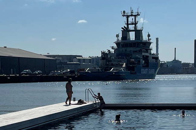 Personer på brygga och i vatten, kustbevakningsfartyg och kranar i hamn.