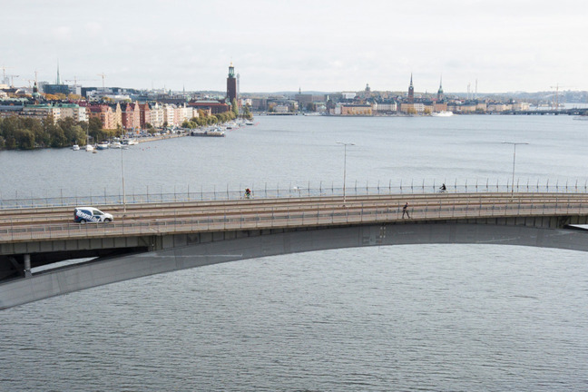 En bro över vatten med stadslandskap i bakgrunden, byggnader och träd med höstfärger.