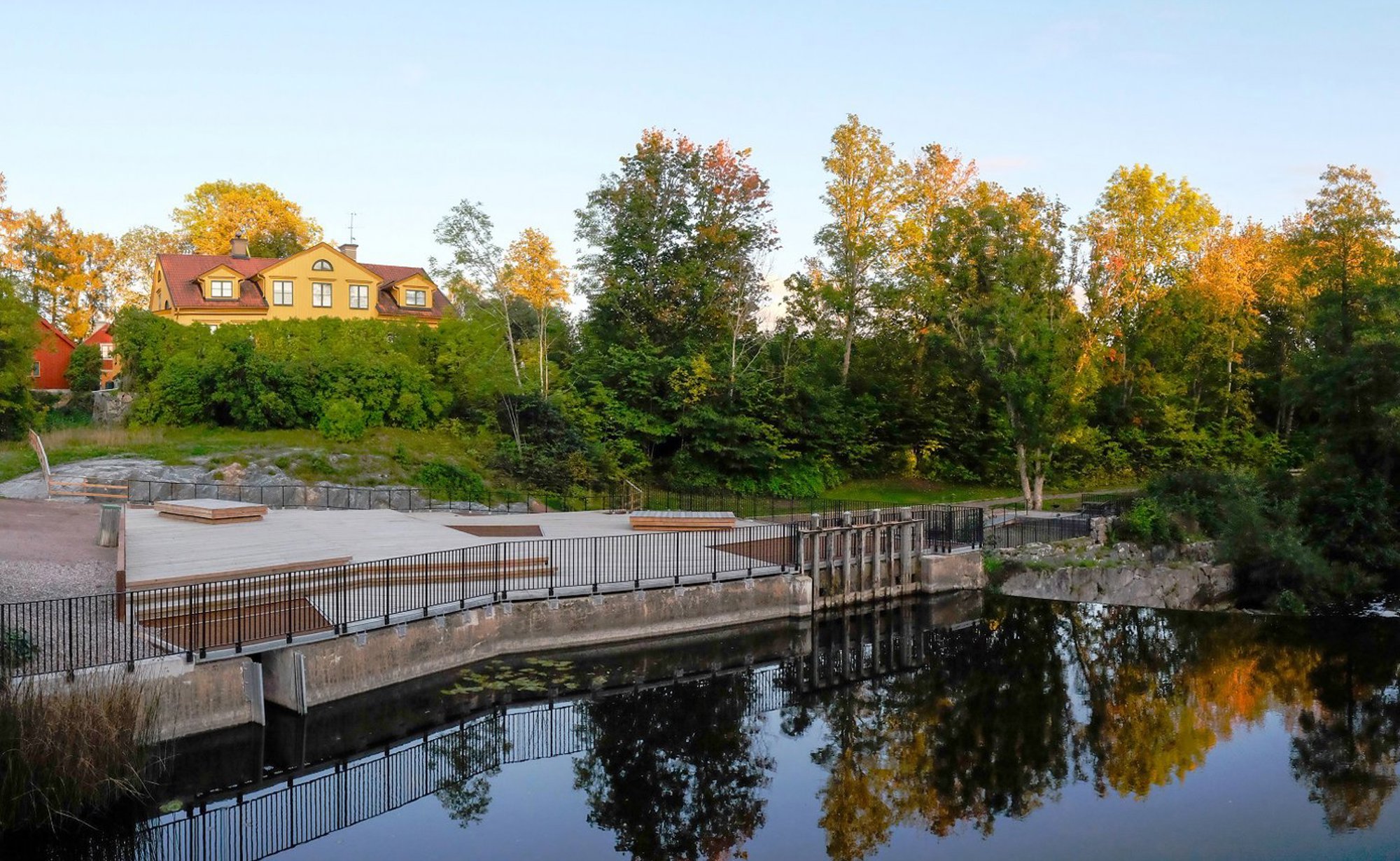 Gul byggnad med röd tak, träd och bro över sjö med spegelbild.