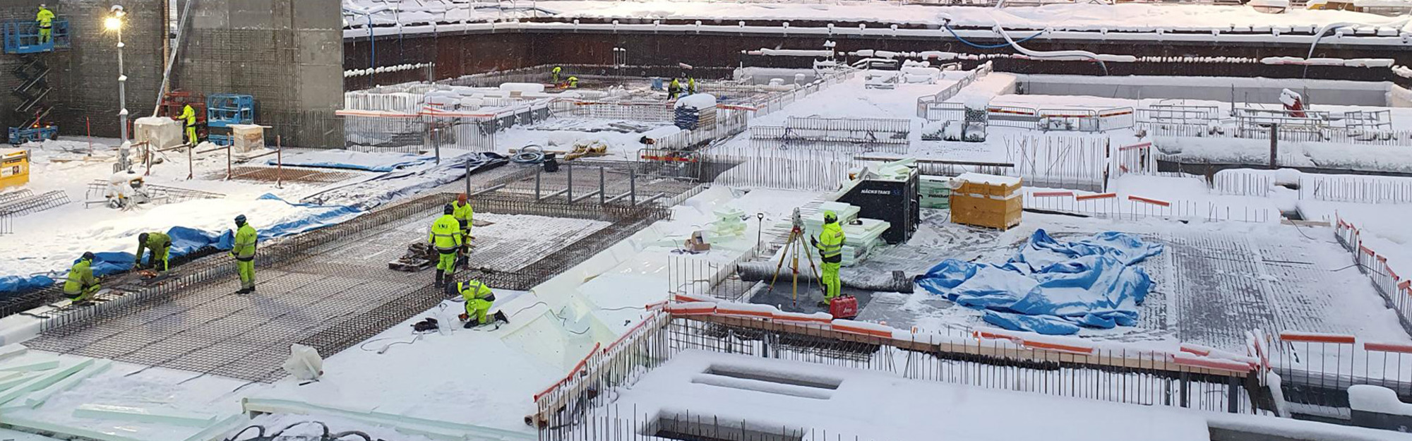 Byggarbetsplats täckt av snö med arbetare i gröna vistrar och byggnadsmaterial.