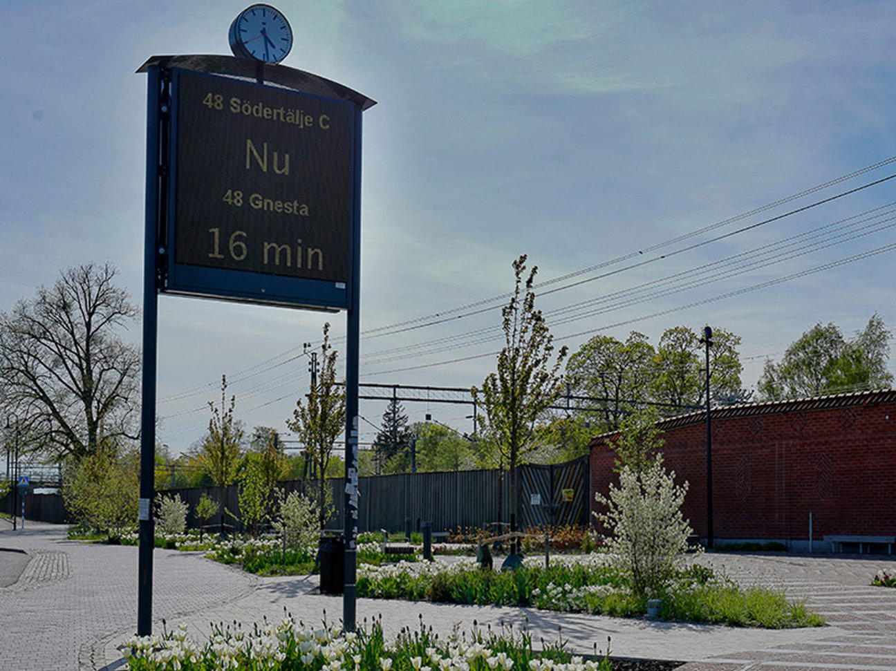 Skylt med avgångstider, klocka, träd och en röd tegelbyggnad i bakgrunden.