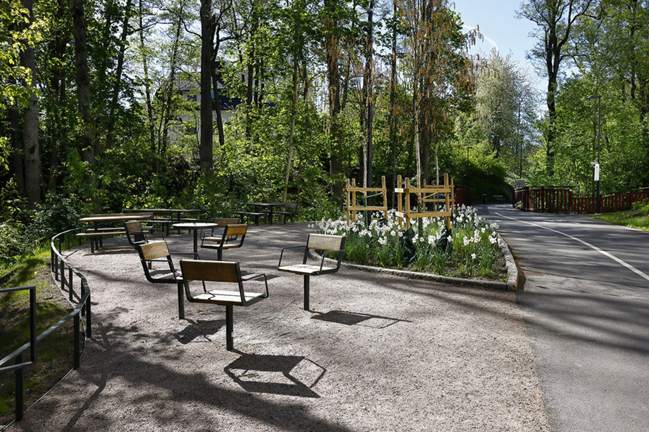 Grusbelagd plats med bänkar, bord och blomsterbädd med vita blommor omgiven av träd och en väg.