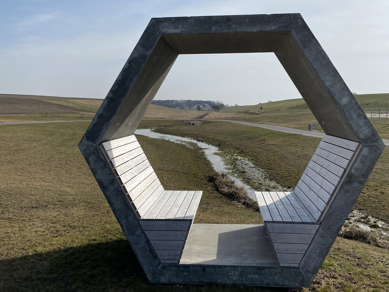 En betongstruktur i form av en sexhörning med bänkar, placerad i ett öppet landskap med en liten vattendrag och vägar i bakgrunden.