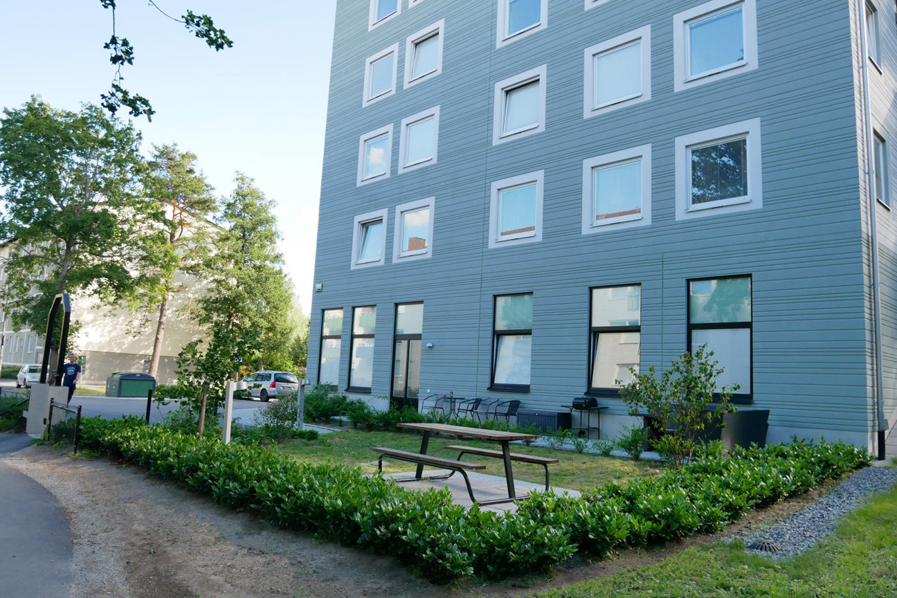 Grå bostadsbyggnad med vita fönster, grönska och picknickbord framför.