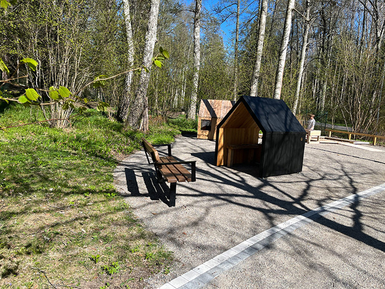 En park med bänk, två trähus och en person i bakgrunden omgiven av träd.