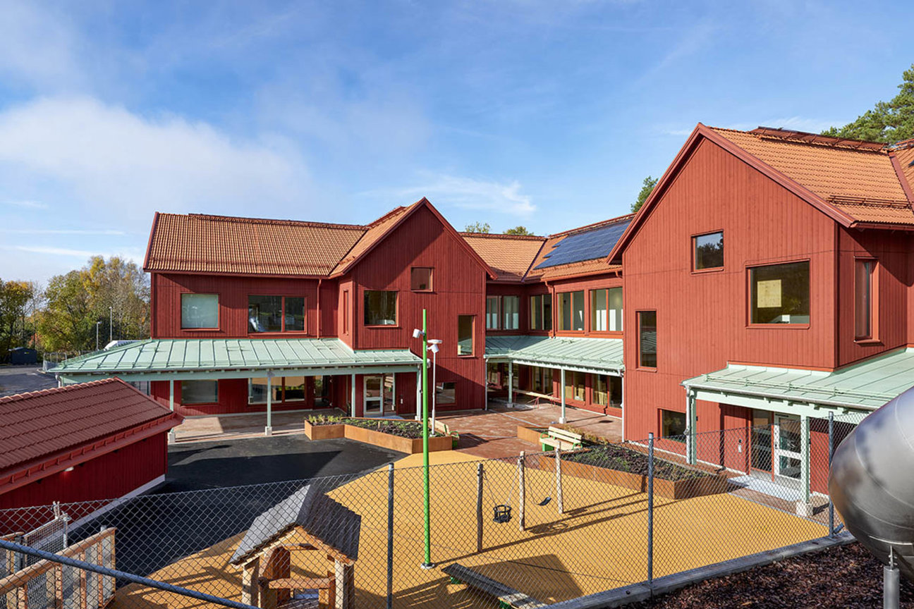 Rött trähuskomplex med lekplats och staket under blå himmel.