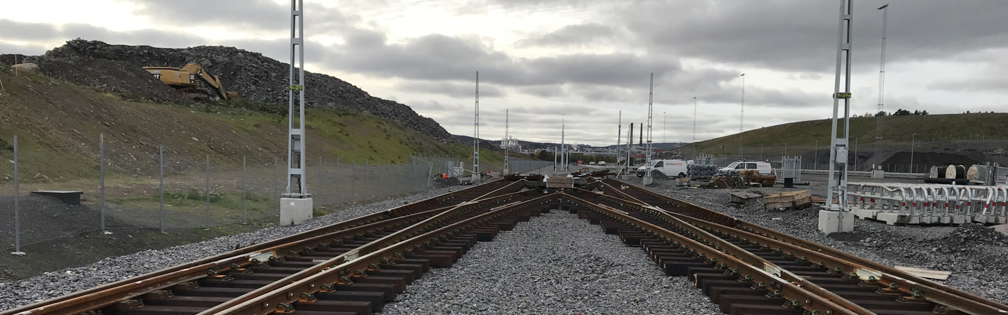 Järnvägsspår som delar sig med byggnadsutrustning och kullar i bakgrunden under molnigt väder.