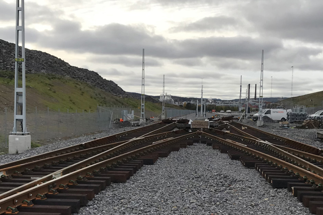 Järnvägsspår som delar sig med byggnadsutrustning och kullar i bakgrunden under molnigt väder.