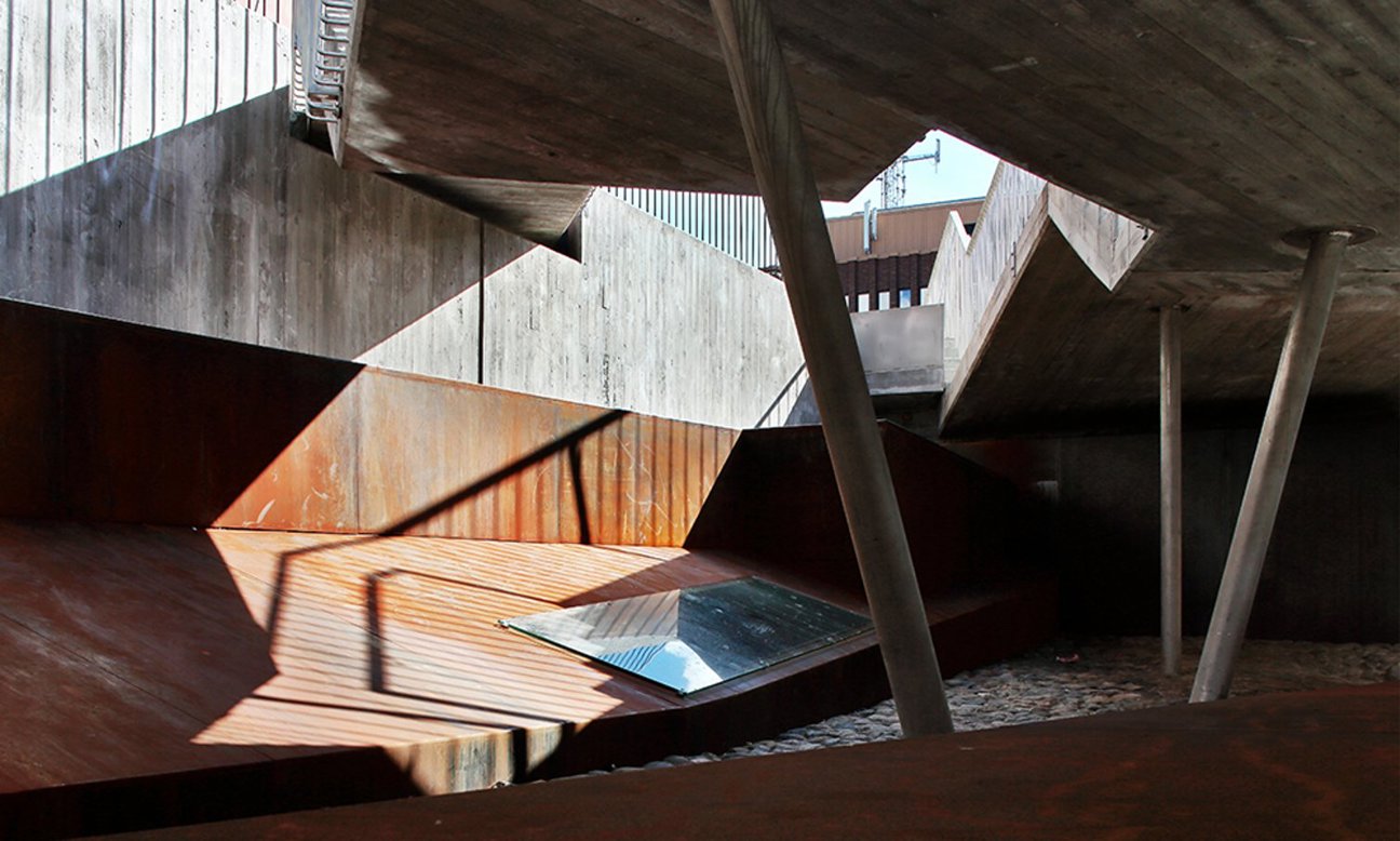 Arkitektoniskt utrymme med betongkonstruktioner, metallstolpar, skuggor och en glaspanel i marken med grusgolv.