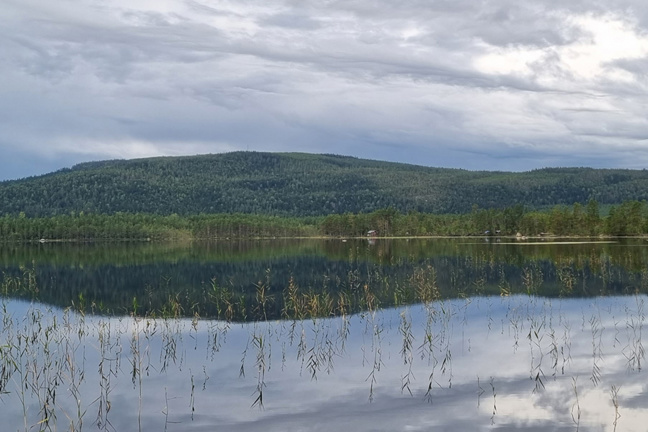 En lugn sjö som speglar molnigt himmel och skogklädda kullar.