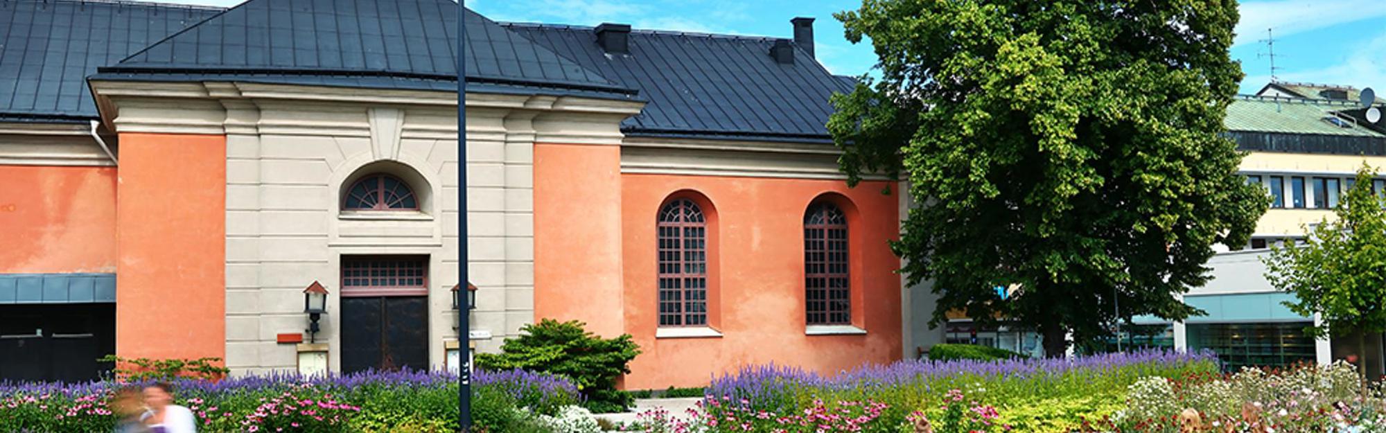 En byggnad med orange och ljusbeige fasad, bågformade fönster, mörkt tak och blommor i förgrunden.