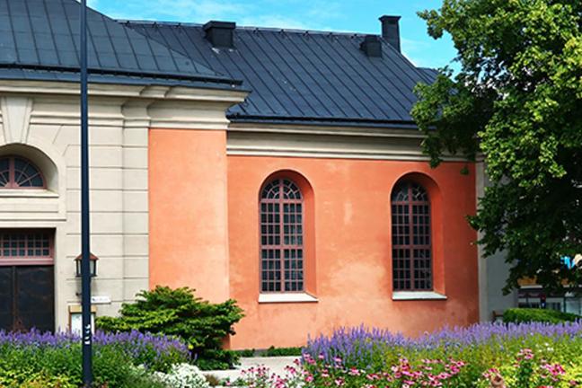 En byggnad med orange och ljusbeige fasad, bågformade fönster, mörkt tak och blommor i förgrunden.