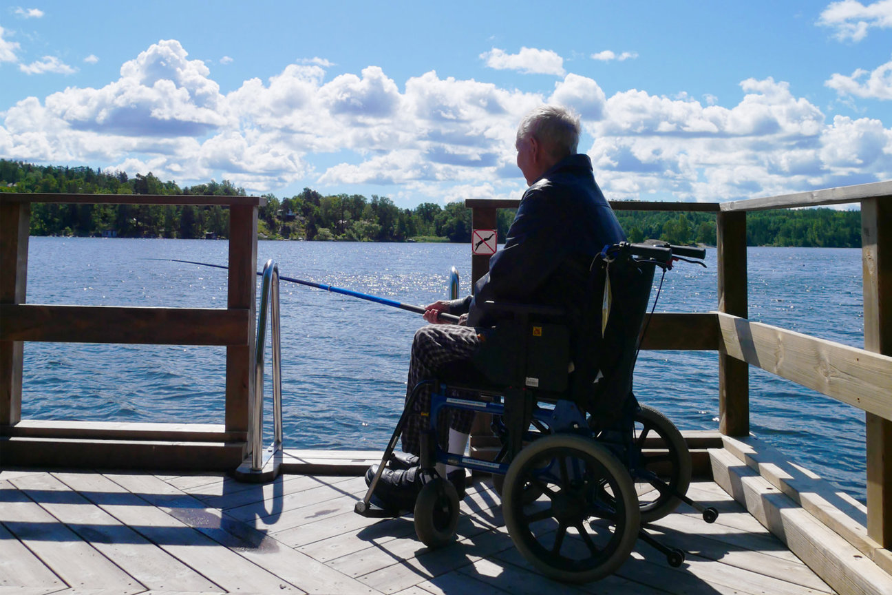 En person i rullstol på en träbrygga vid en sjö med fiskestav. Blå himmel med moln och träd i bakgrunden.