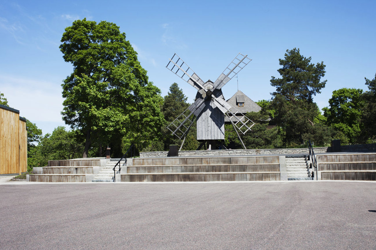 En träväderkvarn med steniga trappor och gröna träd i bakgrunden under klarblå himmel.