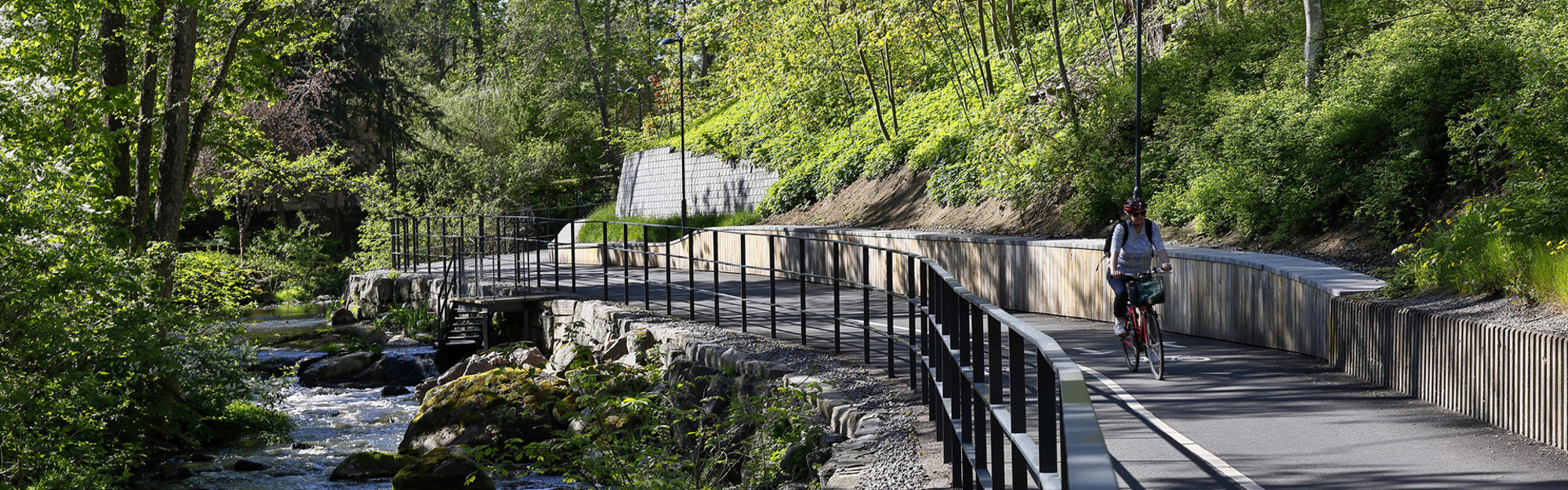 En person cyklar på en cykelbana bredvid ett vattendrag med stenar och grönska i skogsmiljö.